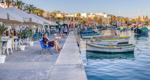 Un paseo marítimo con filas de mesas al aire libre, una hilera de barcos amarrados y edificios al fondo.