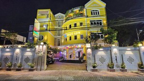 Un hotel muy iluminado con fachada amarilla, una entrada protegida por una reja y plantas en macetas alrededor del perímetro.