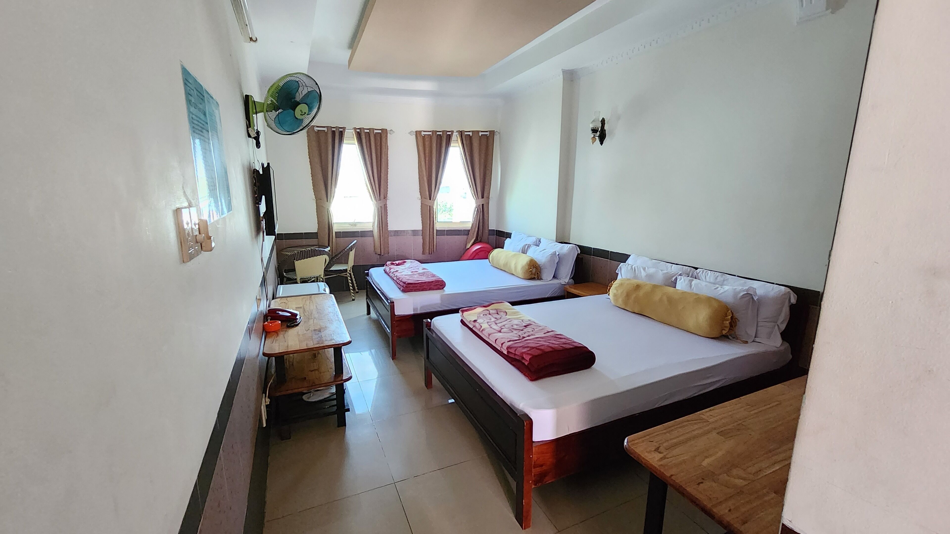 A hotel room with two beds, a small table, a chair, and a fan.