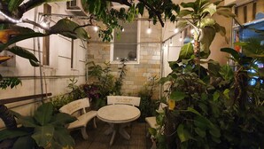 Un espacio al aire libre con una mesa y sillas de piedra, rodeado de abundante vegetación e iluminación colgante.