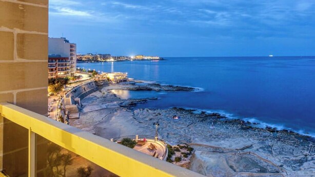 A coastal view from a balcony overlooking a beach with a rocky shoreline, a pier, and buildings in the background.