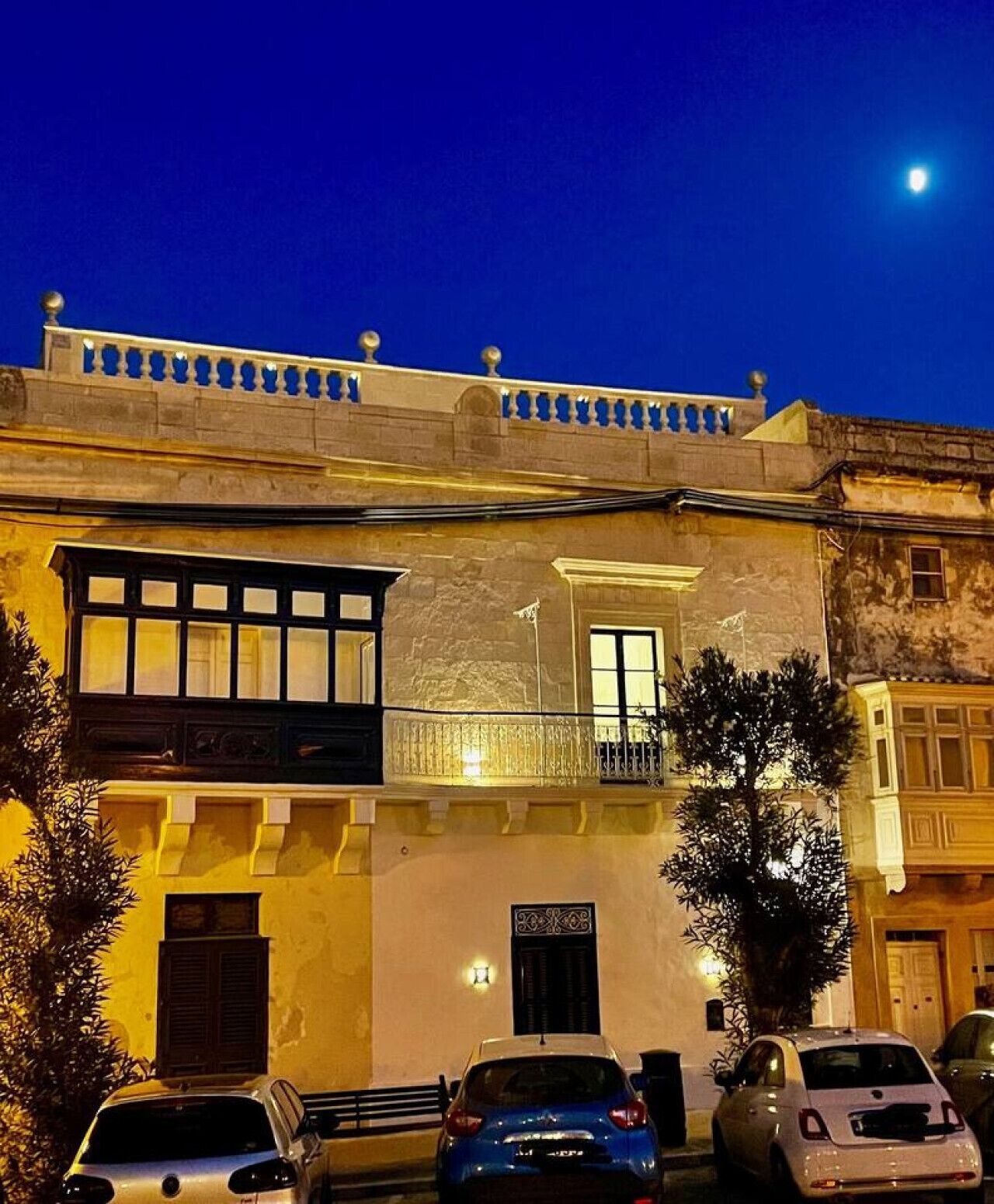 A two-story building with a balcony, a door, and a window. There are cars parked in front.