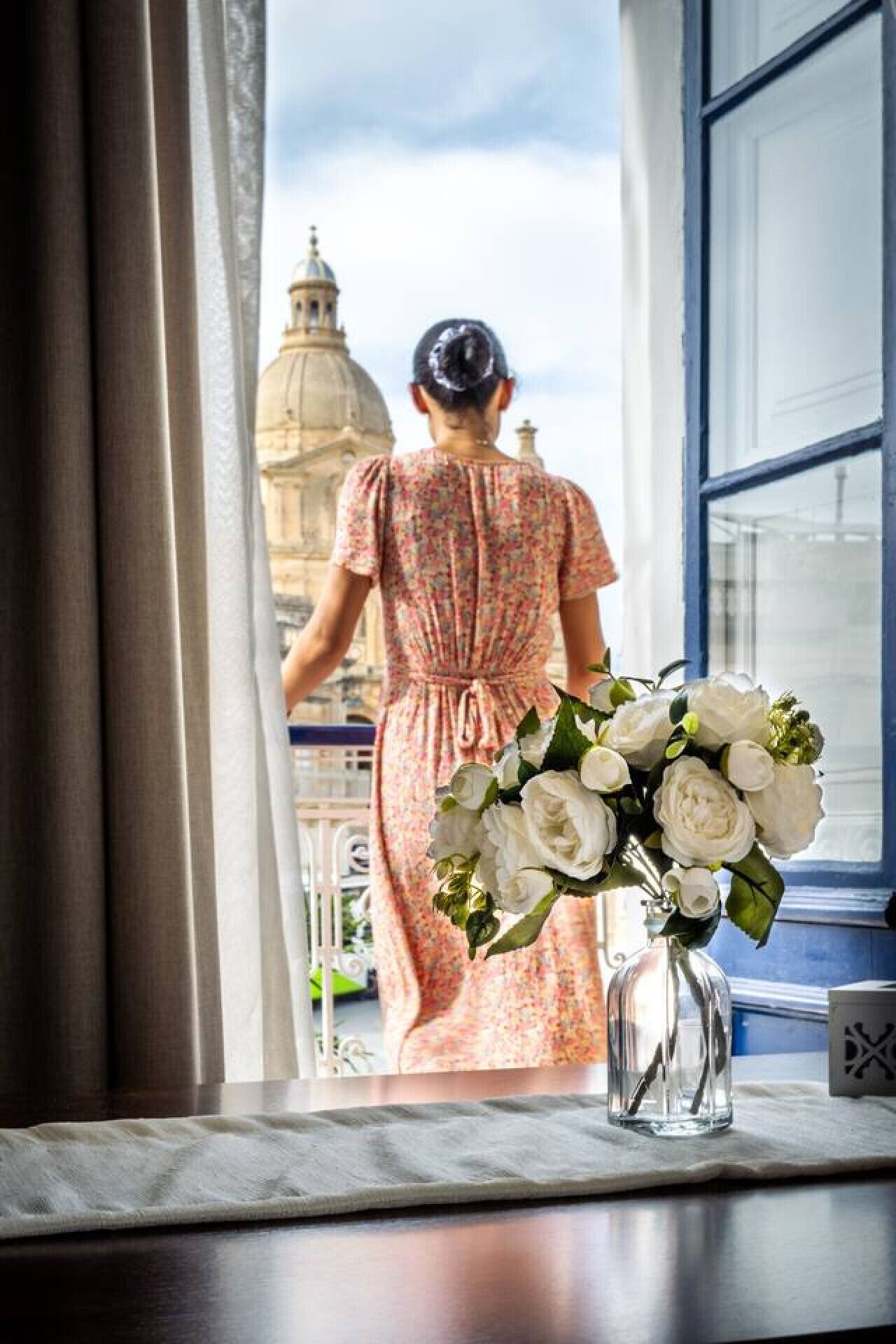 Una habitación con un ventanal grande, un jarrón con flores blancas y vistas a un edificio con cúpula.