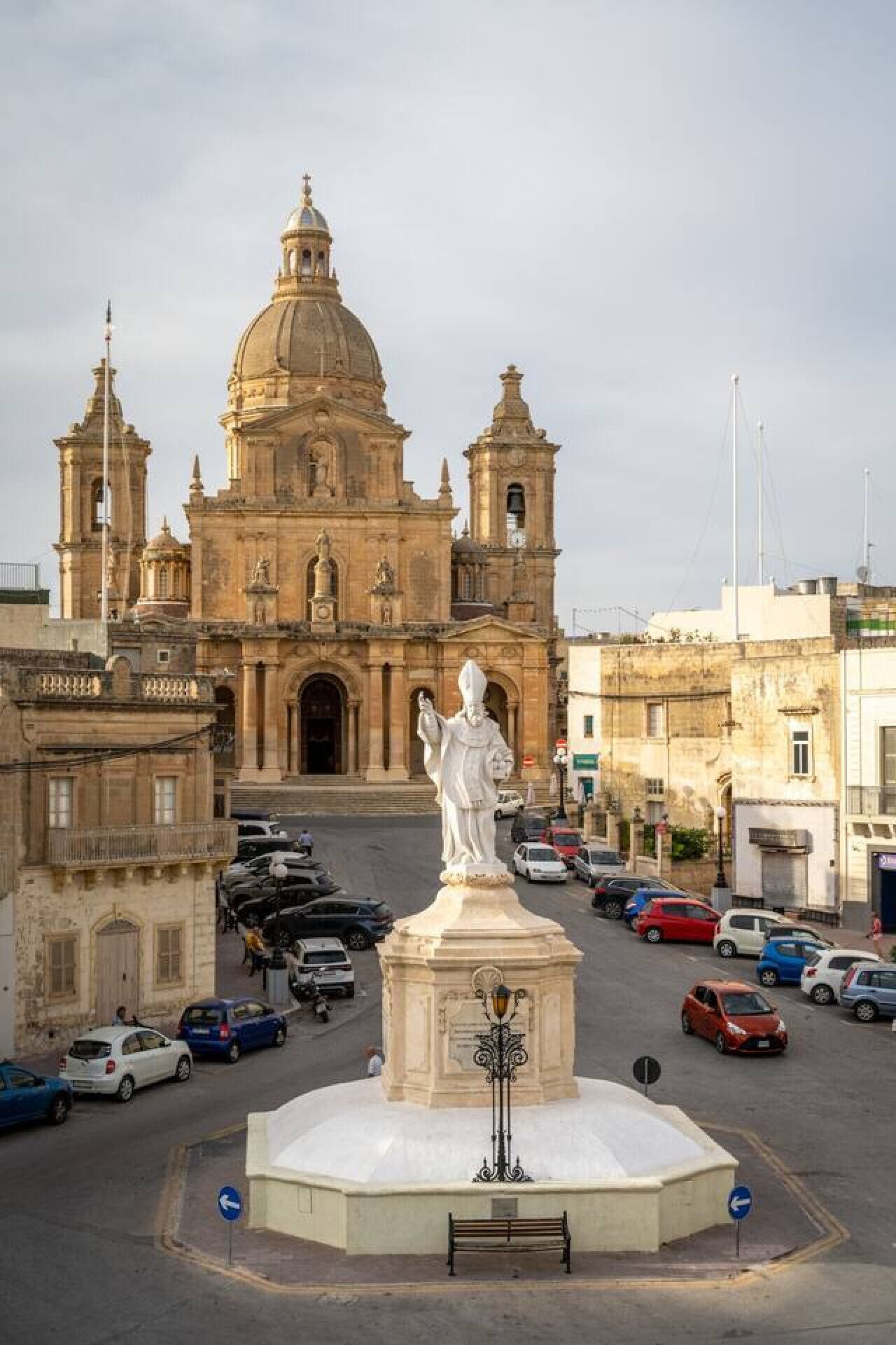 A historic building with a large dome and two bell towers, surrounded by parked cars and a statue in the foreground.