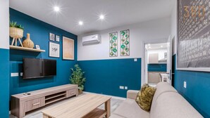 Un salon moderne avec un mur d’accent bleu, un téléviseur à écran plat, une table basse en bois et un canapé beige agrémenté d’un plaid en fausse fourrure.