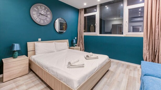 Une chambre à coucher moderne avec un grand lit, deux tables de chevet, une horloge murale, un miroir et une fenêtre avec des rideaux.