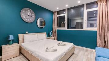 A modern bedroom with a large bed, two bedside tables, a wall clock, a mirror, and a window with curtains.