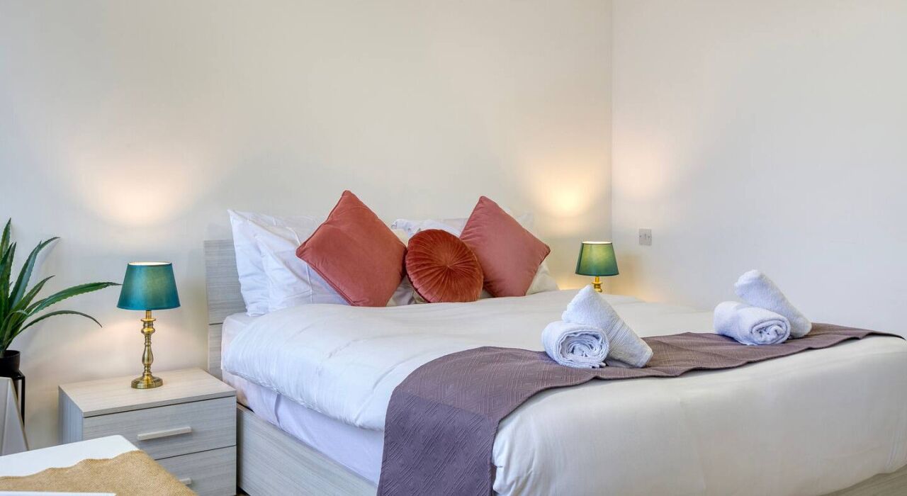A neatly made bed with white linens, a brown throw blanket, and two rolled towels. There are two bedside lamps, one green and one blue, and a potted plant on the left side.