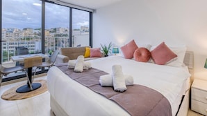 A modern hotel room with a large bed, a seating area with a table and chairs, a view of the city through large windows, and a nightstand with a lamp. - A lovely, studio penthouse with private balcony (San Ġiljan)