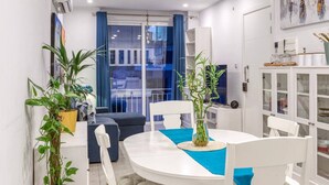 A modern dining area with a round table, white chairs, and blue tablecloths. There are potted plants on the sides and a view of the outside through a glass door.