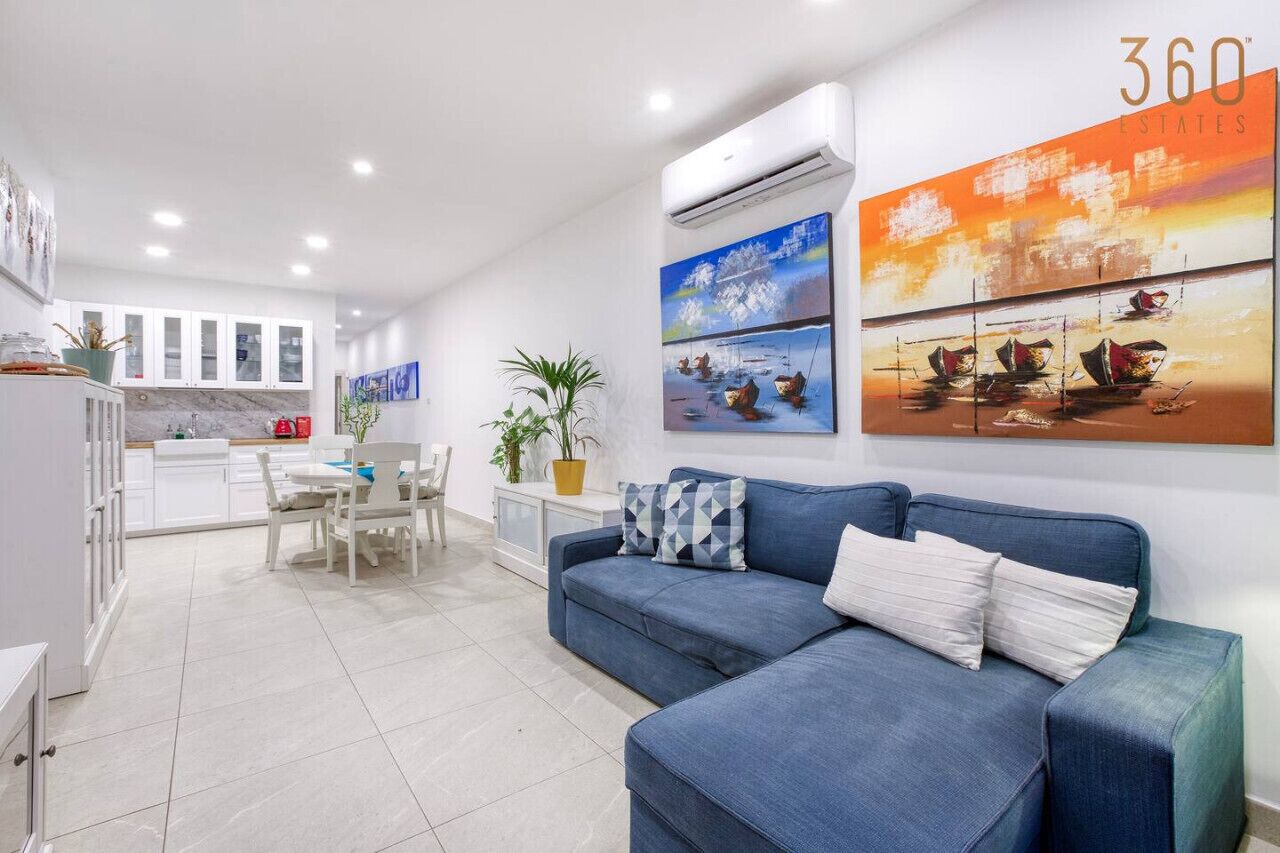 A modern living room with a blue sofa, a dining area with white furniture, and a kitchen in the background.