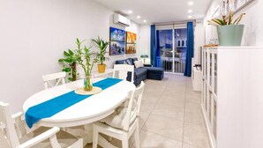 A dining area with a white table, blue runner, and potted plants. There is a sofa and a cabinet in the background.