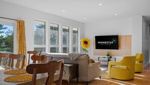 Ein modernes Wohnzimmer mit einer Couch, gelben Sesseln, einem Esstisch mit Platzsets und einem an der Wand befestigten Fernseher.