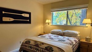 Un dormitorio con una cama, dos lámparas de noche, un ventanal con vistas a árboles y un estante en la pared con dos palas de madera.