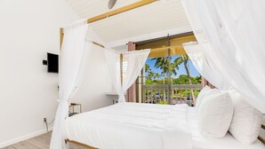 A bed with white bedding and a canopy, a bedside table, a TV mounted on the wall, and a view of palm trees through a window.