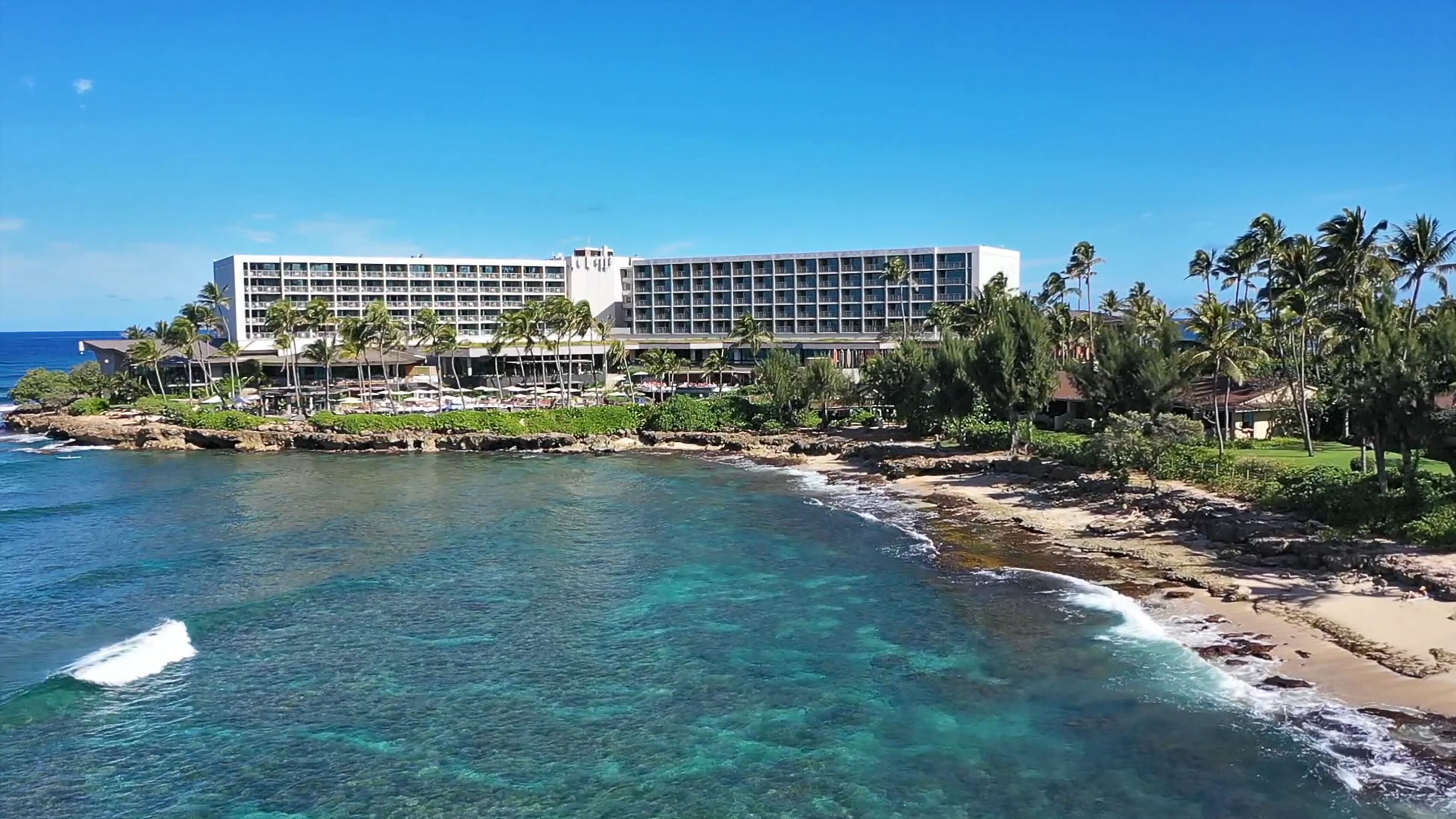 Un hotel frente a la playa con varios pisos, una costa rocosa y palmeras.