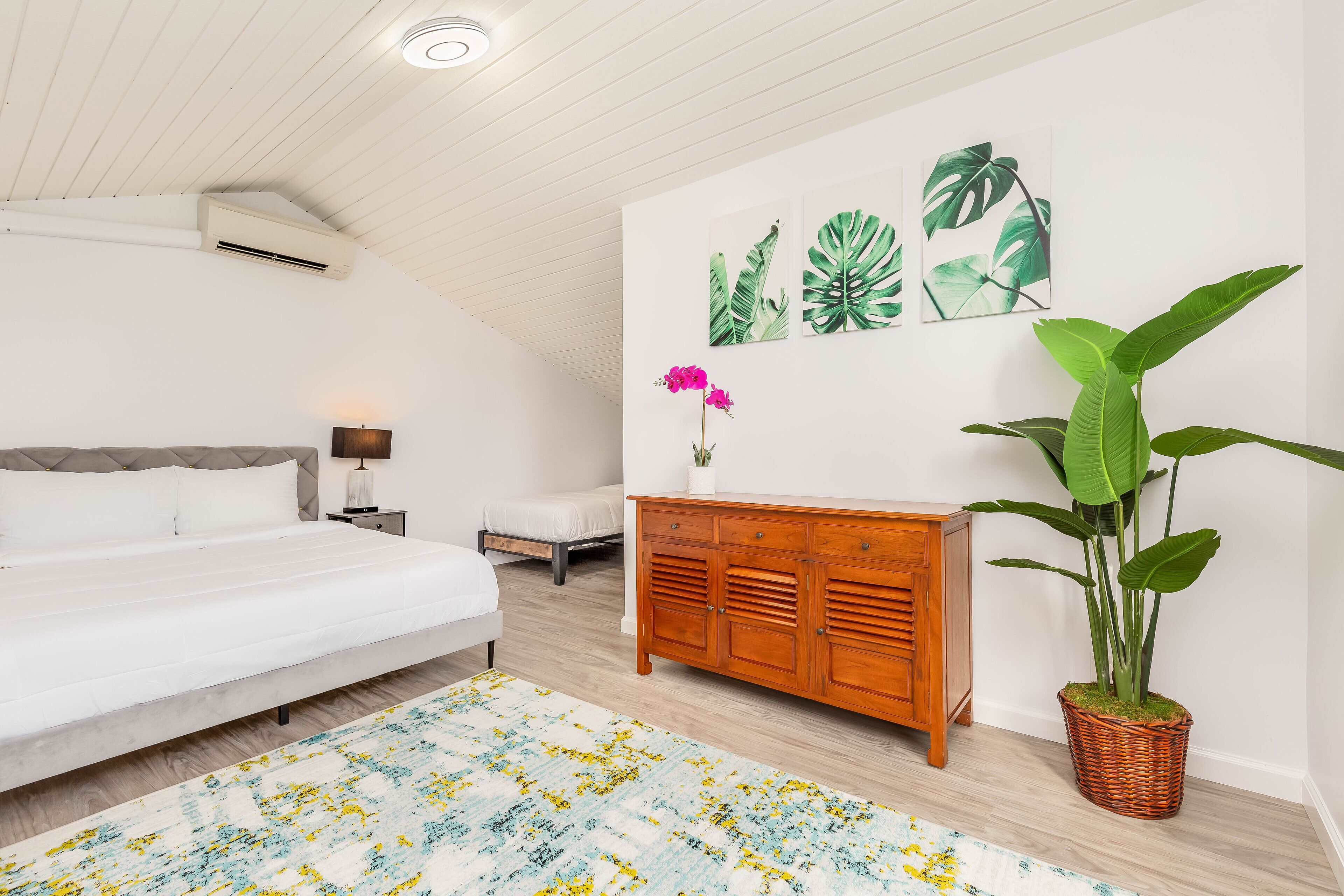 A bedroom with a bed, bedside table, nightstand, dresser, and a large potted plant.