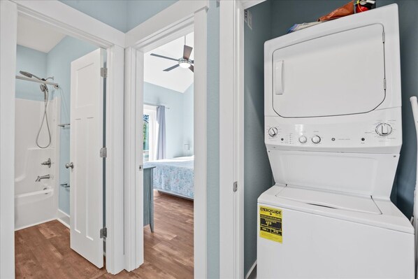 A laundry room with a front-loading washer and dryer, a shower with a glass door, and a hallway leading to a bedroom with a window.