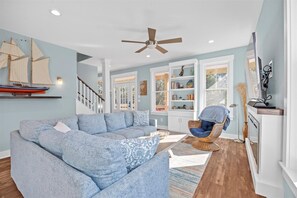 A spacious living room with a large blue sofa, a wicker chair, a ceiling fan, and a bookshelf filled with books and decorative items.
