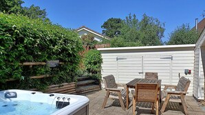 A hot tub with wooden chairs and a table, surrounded by a brick patio and a white fence.