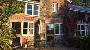 A stone building with a patio area featuring a table and chairs, a door labeled "Wisteria Cottage," and a small window with a curtain. - Little Dunley - Wisteria Cottage (Bovey Tracey)
