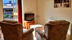 A living room with two floral armchairs, a television on a wooden stand, and a large window with a red curtain. - Little Dunley - Virginia Cottage (Bovey Tracey)