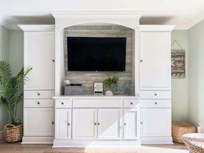 A living room with a large flat-screen TV mounted on a white cabinet, surrounded by white cabinetry, a wicker basket, and a potted plant.