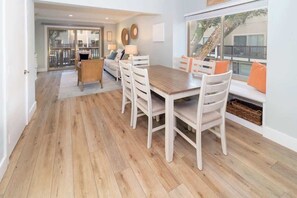Un coin repas avec une table en bois et des chaises blanches, un canapé avec des coussins et une vue par la fenêtre.