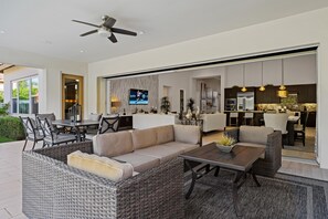 A spacious living area with a sofa, coffee table, and dining table. A television is mounted on the wall, and there is a kitchen area visible through an open door.