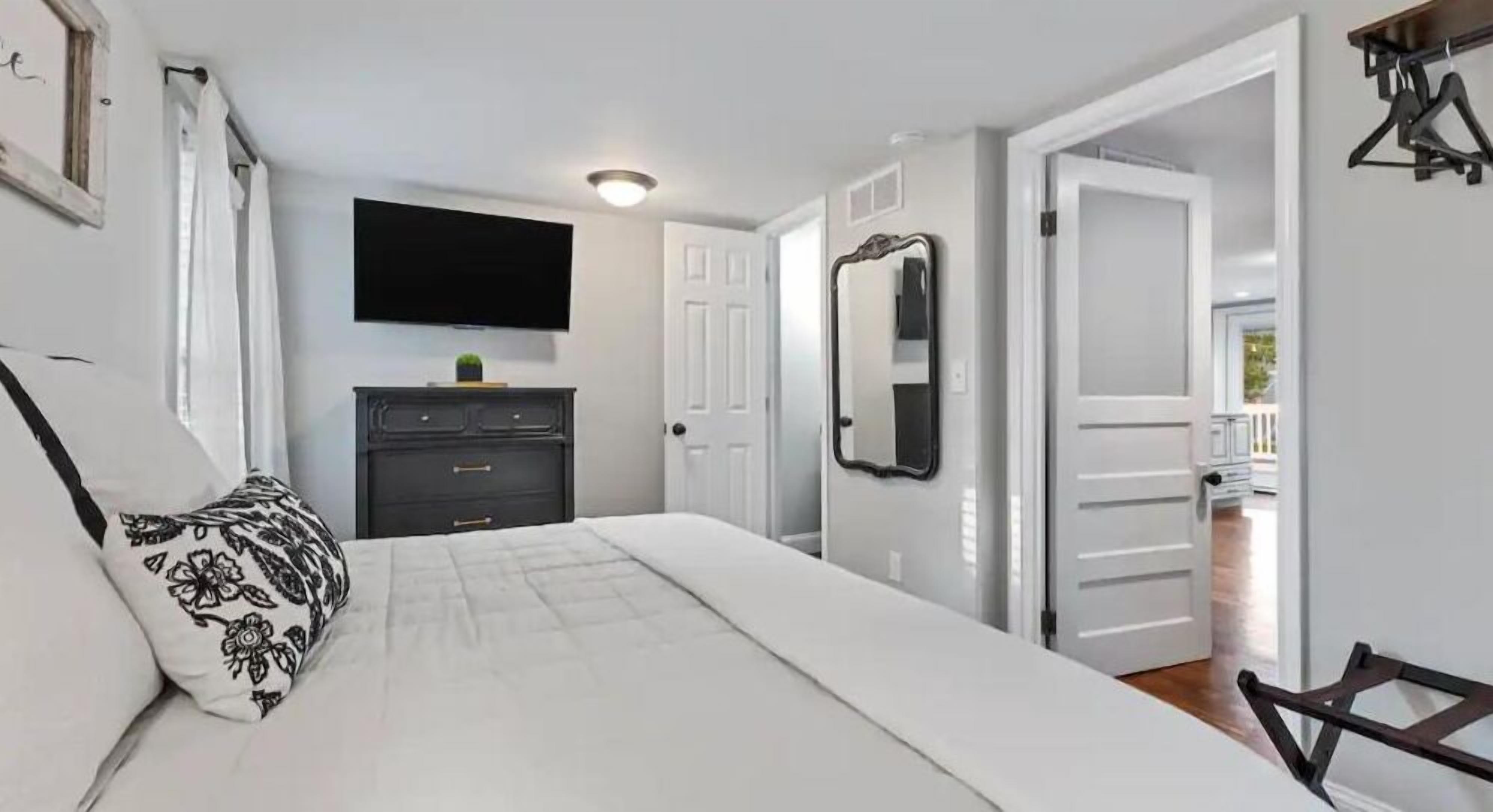 Un dormitorio con cama, cómoda, televisor, espejo y una puerta que da a otra habitación.