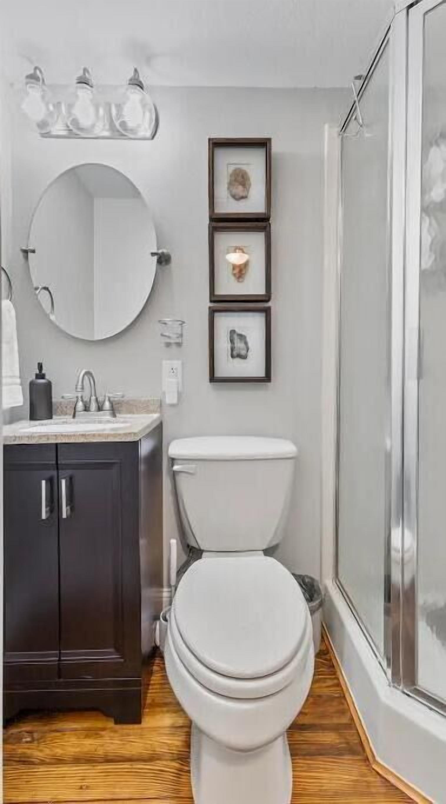 A modern bathroom with a toilet, sink, and mirror. There are framed pictures on the wall and a shower area visible.