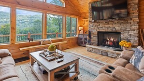 A cozy cabin interior with a stone fireplace, wooden ceiling, large windows overlooking a forest, and a comfortable seating area. - Black Bear Ridge! Newly Built! 4BD/3.5 BA (Maggie Valley)