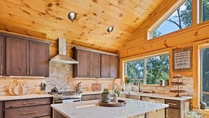 A modern kitchen with wooden cabinets, a stone island, and a large window offering a view of the outdoors. - Black Bear Ridge! Newly Built! 4BD/3.5 BA (Maggie Valley)