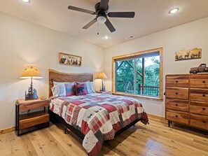 A bedroom with a wooden bed, two bedside tables with lamps, a wooden dresser, a window with a view of trees, and a ceiling fan. - Black Bear Ridge! Newly Built! 4BD/3.5 BA (Maggie Valley)