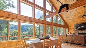 A wooden cabin with a dining table and chairs, large windows, and a stone fireplace. - Black Bear Ridge! Newly Built! 4BD/3.5 BA (Maggie Valley)