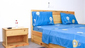 Une chambre à coucher avec un lit en bois, une literie bleue ornée de motifs de tournesols, et une petite table de chevet en bois sur laquelle se trouvent une télécommande et une bouteille.