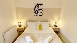 Un lit bien fait, avec une couette blanche matelassée, deux serviettes enroulées et un seul oreiller jaune. On peut voir deux lampes de chevet sur des tables de nuit en bois, et un radiateur est visible sur la gauche.