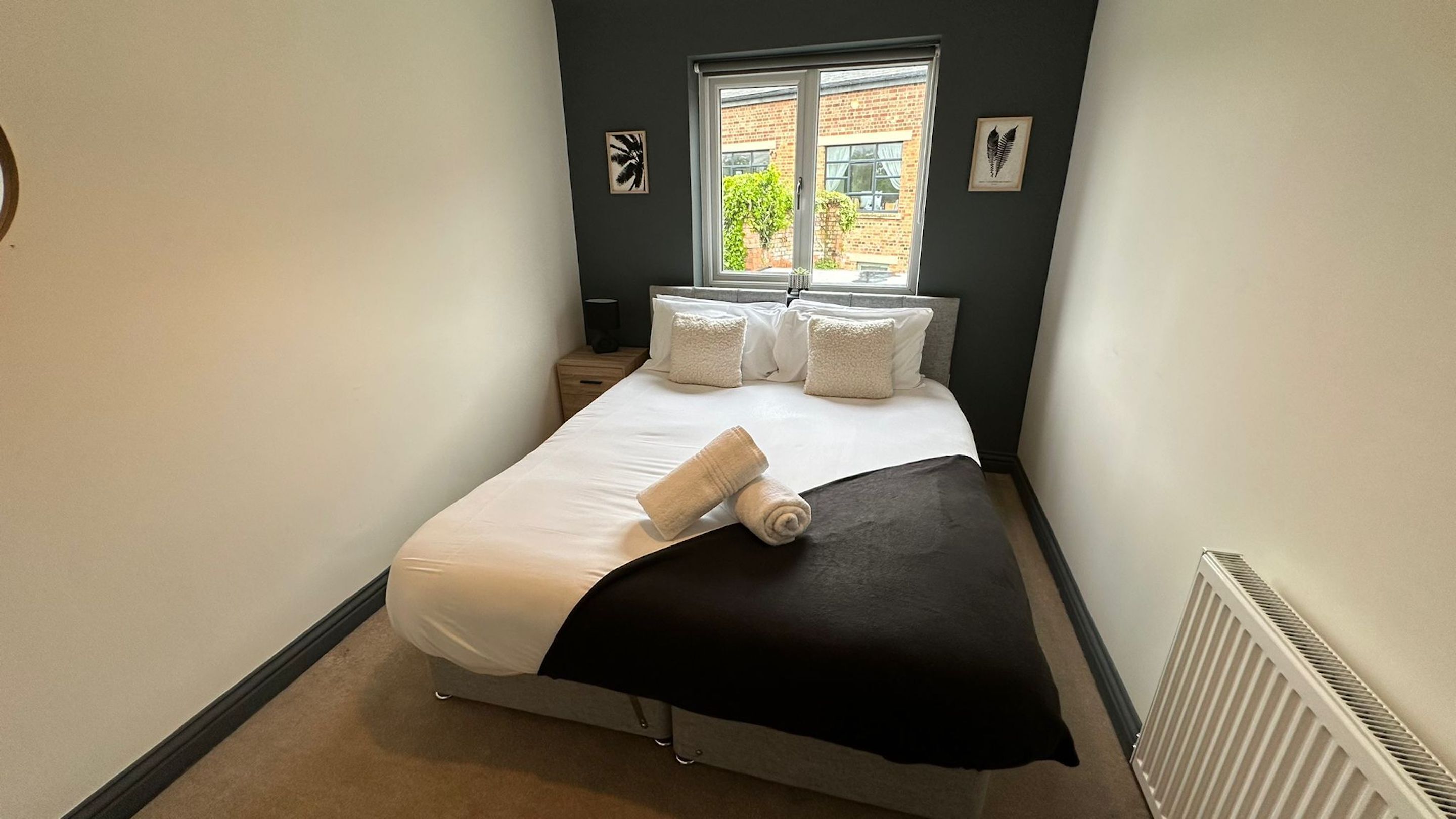 A small, neatly made bed with white linens and a black throw, a window with a view of a brick building, and two framed botanical prints on the wall.