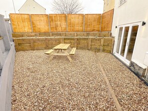 Un jardin avec une table de pique-nique en bois entourée de gravier, une clôture en bois et un bâtiment doté d’une fenêtre.