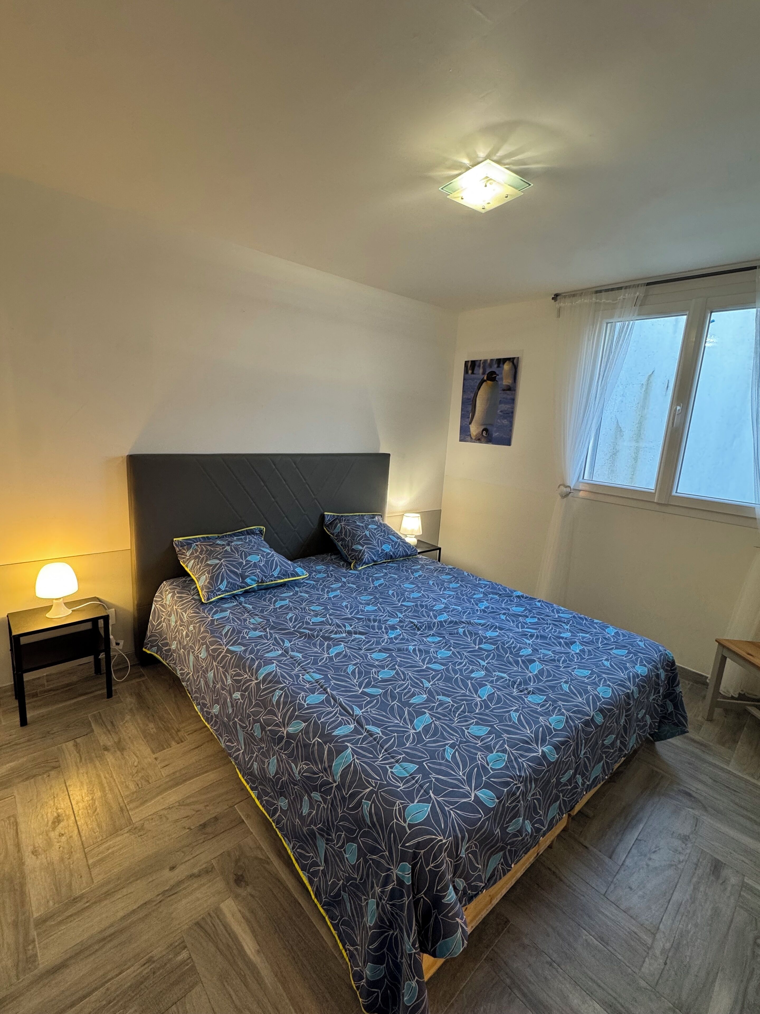Een slaapkamer met een bed, twee nachtkastjes, een nachtlampje, plafondverlichting, een raam met gordijnen en een aan de muur hangend schilderij.