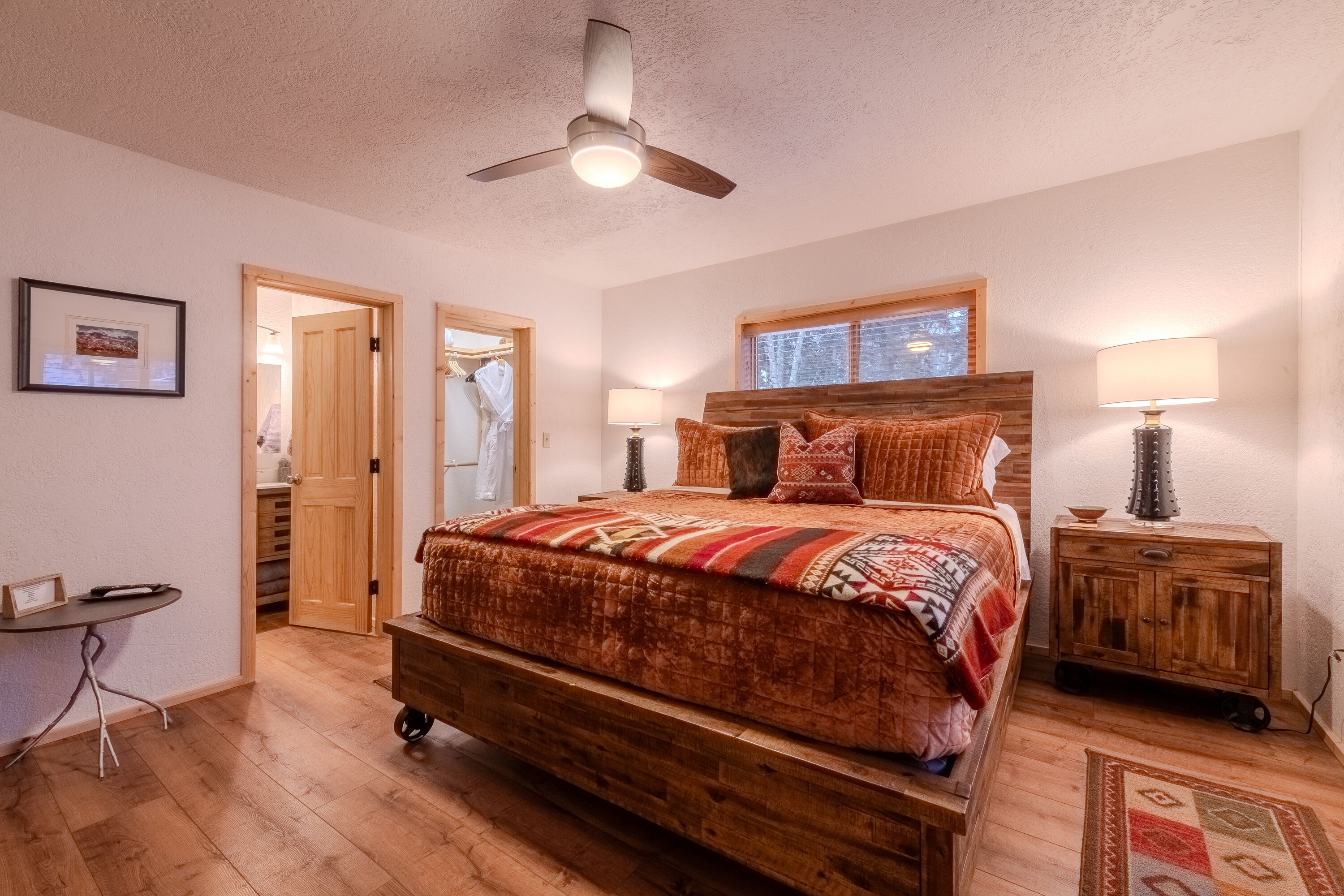 Un dormitorio con una cama de madera, un ventilador de techo, una mesita de noche con lámpara y suelo de madera.