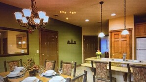 Un comedor con una mesa de madera dispuesta para cuatro personas, rodeada de sillas. Hay una araña sobre la mesa y luces colgantes sobre la zona del bar.