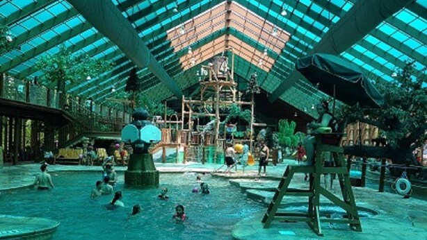 Parque acuático interior con una amplia piscina central, múltiples zonas de juegos y un techo de cristal alto.