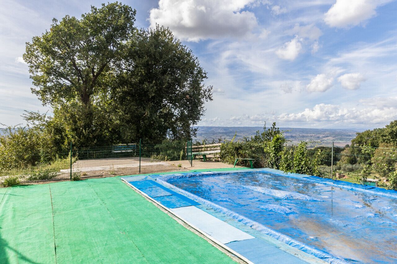 Ein Swimmingpool mit grüner Liegefläche und blauer Wasseroberfläche, umgeben von Bäumen und einem Zaun, mit Blick auf entfernte Hügel.