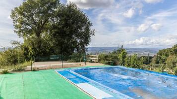 Ein Swimmingpool mit grüner Liegefläche und blauer Wasseroberfläche, umgeben von Bäumen und einem Zaun, mit Blick auf entfernte Hügel.