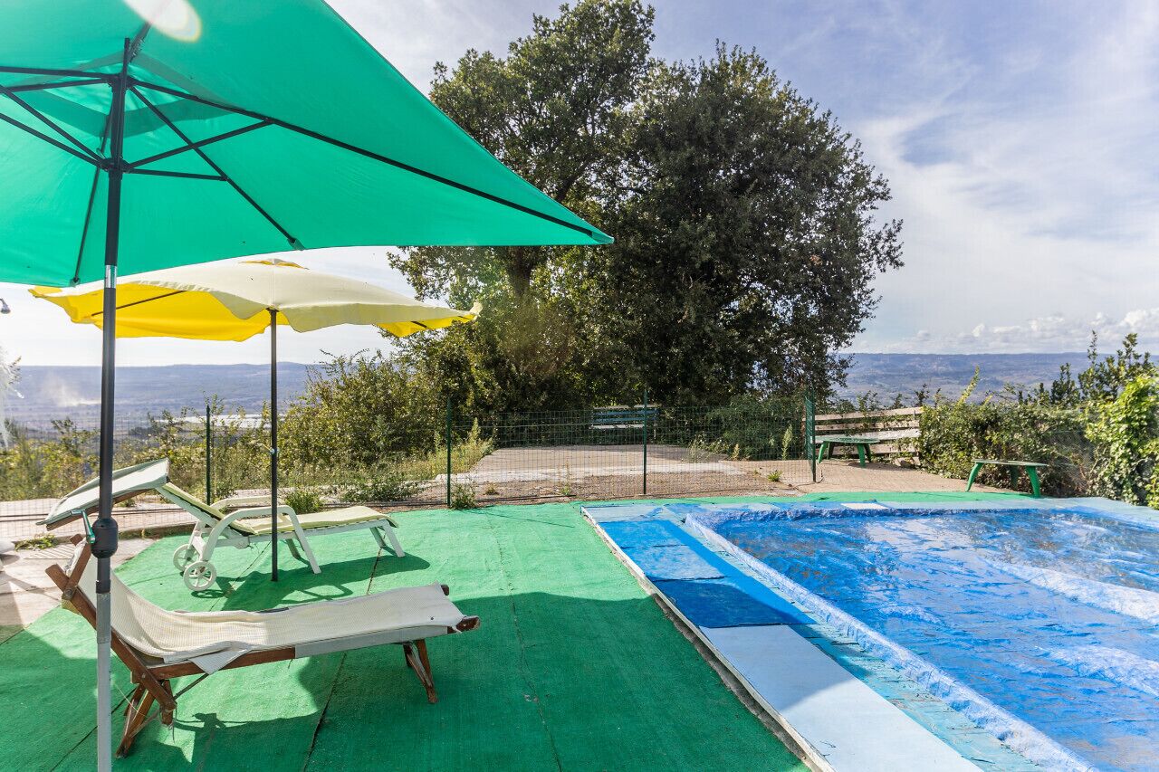 Ein Schwimmbecken mit grüner Liegefläche und Liegestühlen, umgeben von Bäumen und Blick auf die Landschaft.