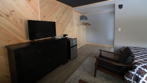 Ein Schlafzimmer mit Holzwand, einem schwarzen Schminktisch, einem Fernseher, einem Bett mit gemusterten Betttextilien und einem Sessel.
