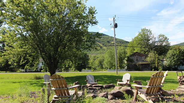 Ein ländlicher Außenbereich mit Adirondack-Stühlen und einem großen Baum, vor einer Kulisse aus einem grünen Feld und einem Haus auf einem Hügel.