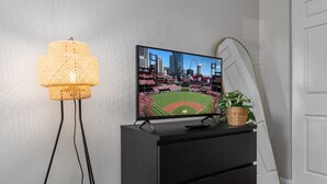 Un téléviseur posé sur une commode noire, affichant une image d’un terrain de sport au cœur d’une ville, une lampe en osier et une plante.