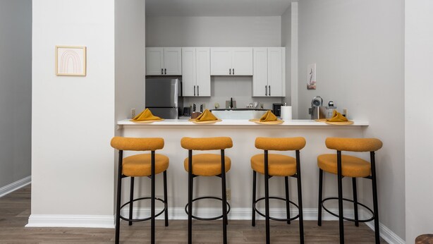 Une cuisine moderne avec des meubles blancs, un réfrigérateur en acier inoxydable et quatre tabourets de bar dotés de coussins jaunes.